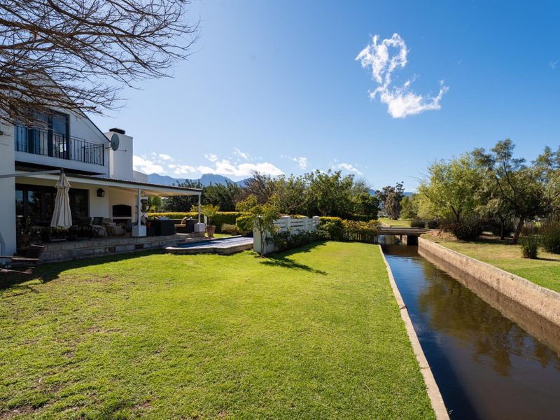 Rear view of the property featuring a covered patio with built-in braai, swimming pool, and a landscaped garden bordering a scenic canal.