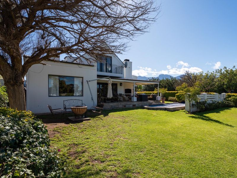 Rear view of the property featuring a spacious lawn, covered patio with built-in braai, swimming pool, and an upper-level balcony.