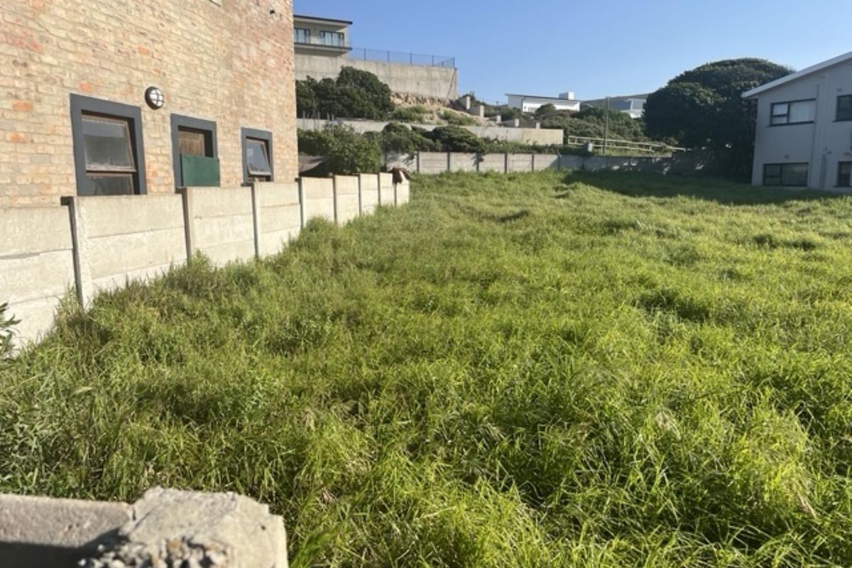 A vacant residential plot featuring overgrown grass, enclosed by a precast concrete boundary wall adjacent to an existing brick building.