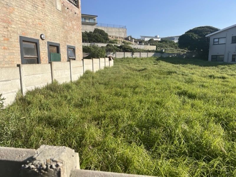 A vacant residential plot featuring overgrown grass, enclosed by a precast concrete boundary wall adjacent to an existing brick building.