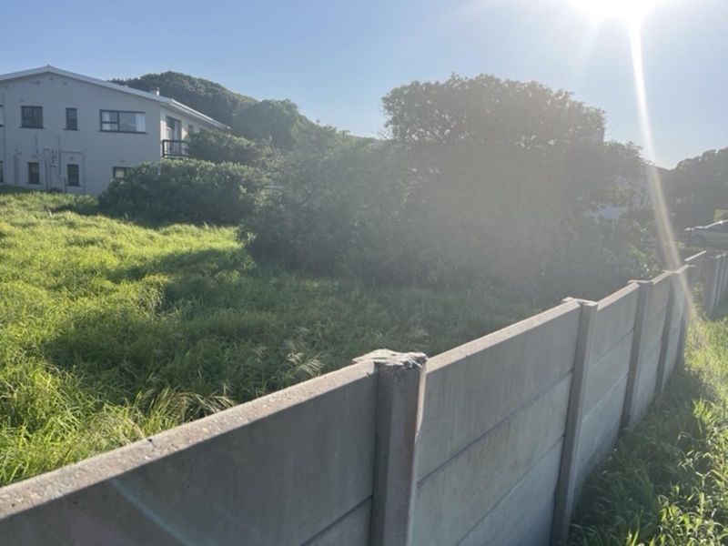A view of a vacant residential plot featuring a precast concrete boundary wall and overgrown grass, with neighboring residential structures visible in the background.