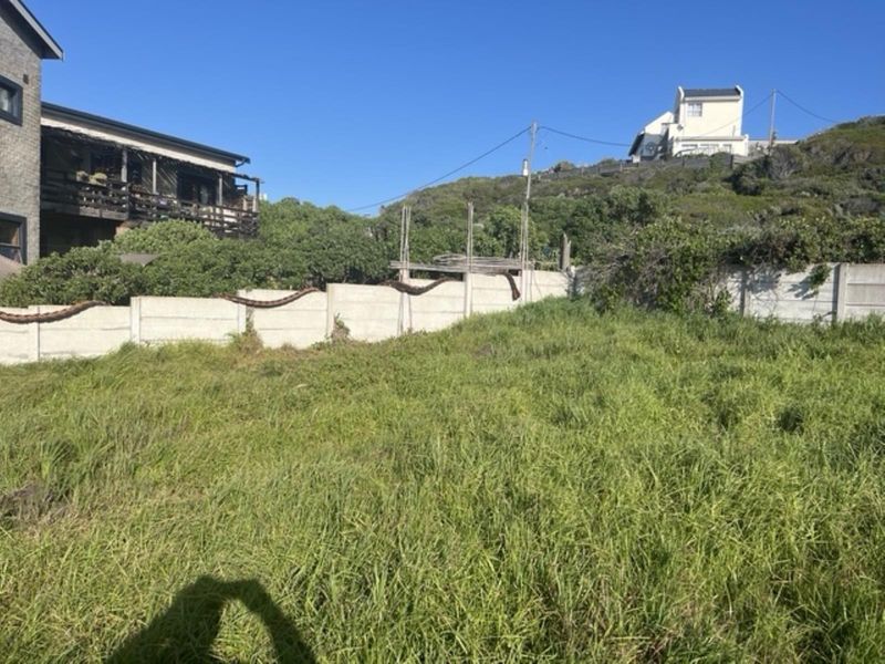 A vacant residential plot featuring overgrown grass and a precast concrete boundary wall, situated in a sloped coastal or hillside neighborhood.
