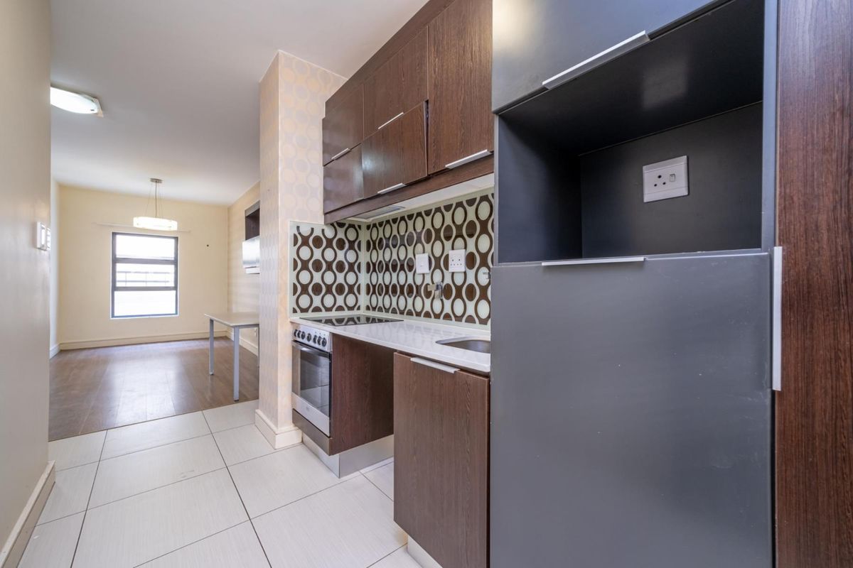 Compact kitchen featuring dark wood cabinetry, built-in oven and hob, and a patterned tiled backsplash, flowing into an open-plan living area.