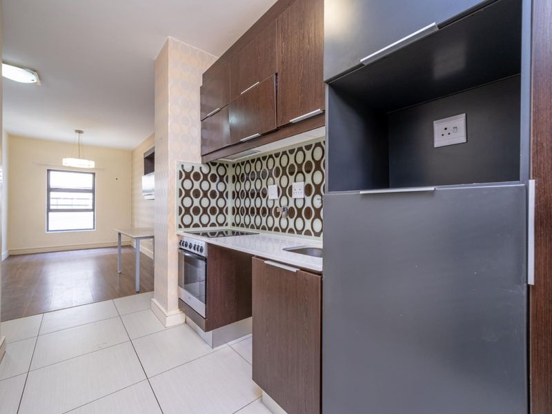 Compact kitchen featuring dark wood cabinetry, built-in oven and hob, and a patterned tiled backsplash, flowing into an open-plan living area.