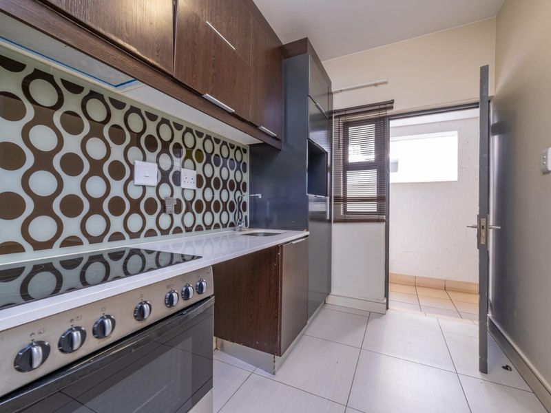 A compact kitchen featuring dark wood cabinetry, a built-in electric stove and oven, patterned splashback, and tiled flooring with direct access to an outdoor area.
