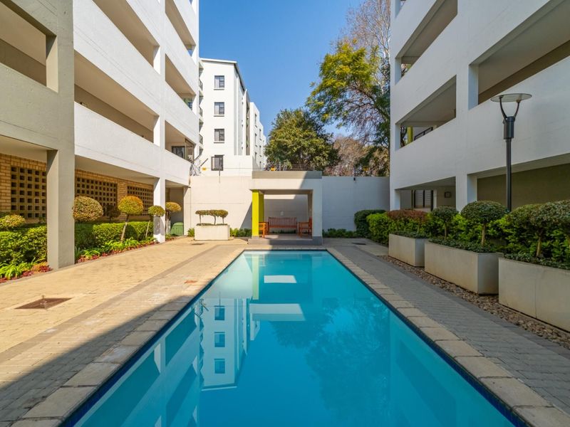 A well-maintained communal swimming pool area situated between apartment blocks, featuring paved surrounds, built-in planters, and a covered seating alcove.