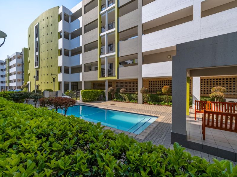 A well-maintained communal swimming pool area featuring a paved deck, landscaped gardens, and a covered patio space within an apartment complex.