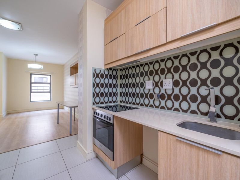 Modern open-plan kitchen featuring light wood cabinetry, integrated electric oven and hob, and a stylish patterned glass splashback, flowing into a bright living area.