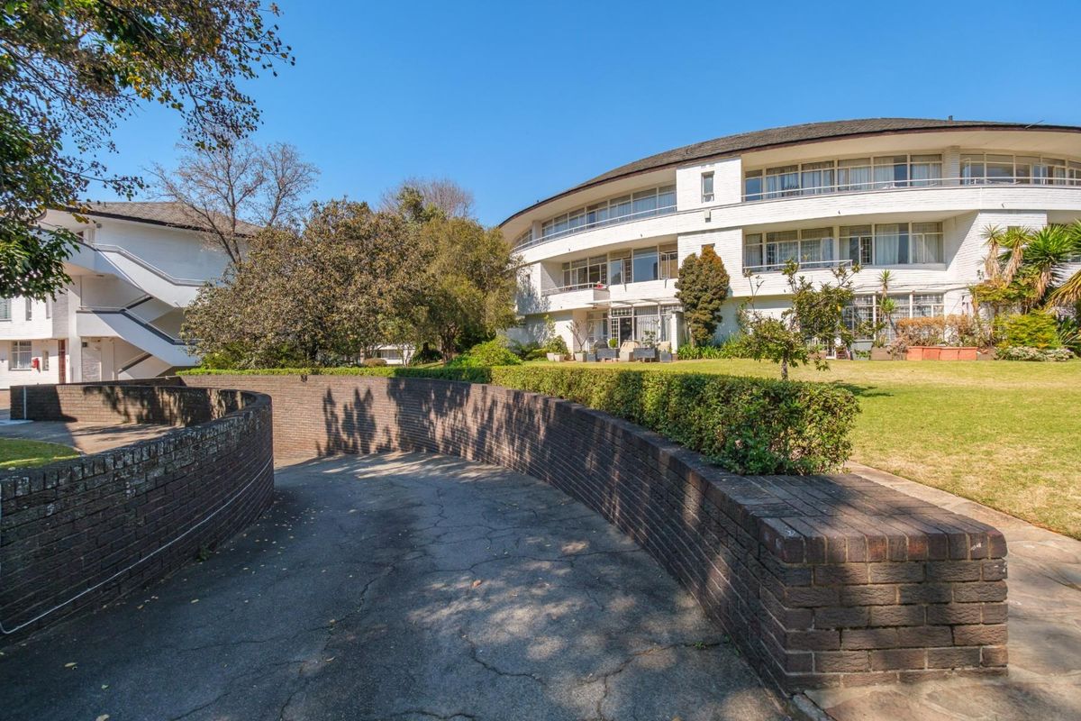 Rear view of the apartment complex featuring a curved building design, landscaped garden areas, and a paved driveway with brick retaining walls.
