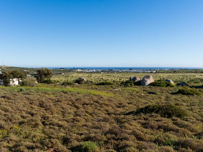 A wide-angle view of a vacant residential plot featuring natural coastal vegetation, large rock formations, and distant ocean views.
