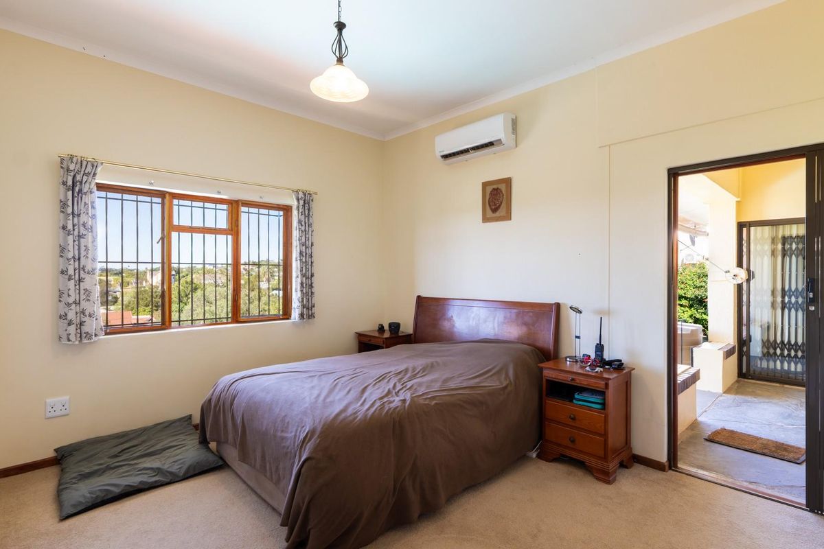 A well-lit bedroom featuring built-in cupboards, an air conditioning unit, and a window with security bars, offering direct access to an outdoor patio area.