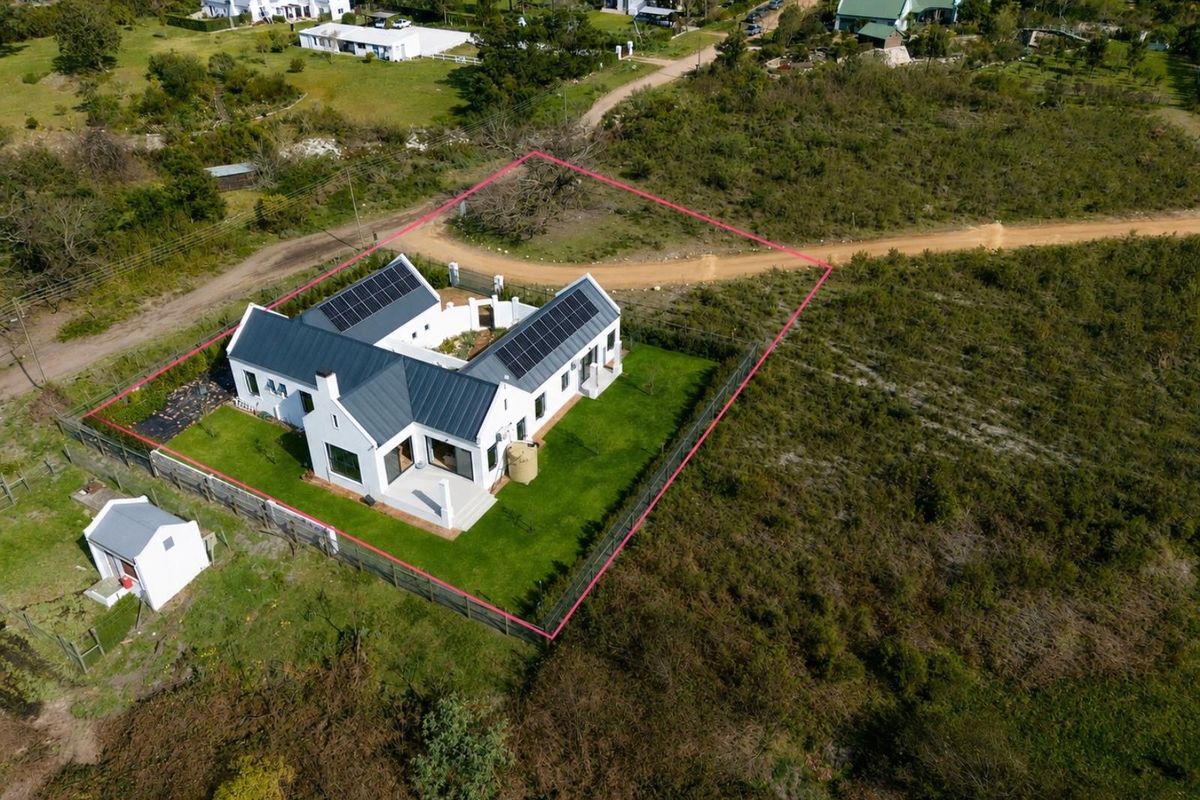 Aerial view of a modern white house with a metal roof, solar panels, and a water tank, situated on a fenced plot.