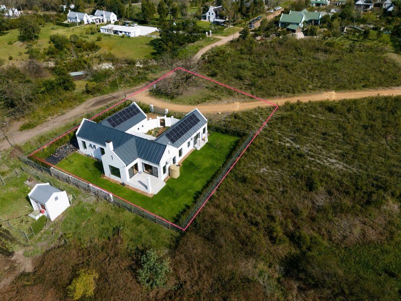 Aerial view of a modern white house with a metal roof, solar panels, and a water tank, situated on a fenced plot.