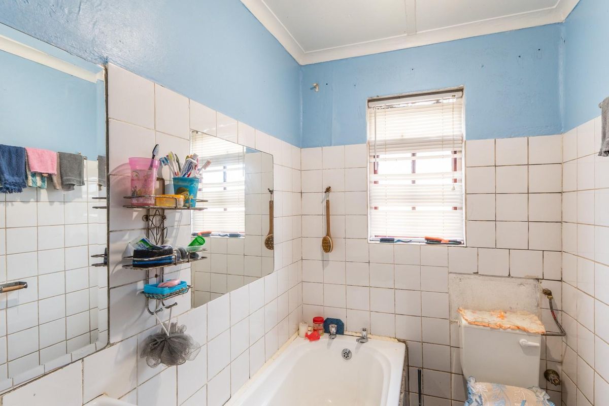 A dated bathroom featuring a built-in bathtub, toilet, and wall-mounted mirror, with ceramic tiling and blue painted walls requiring cosmetic updates.