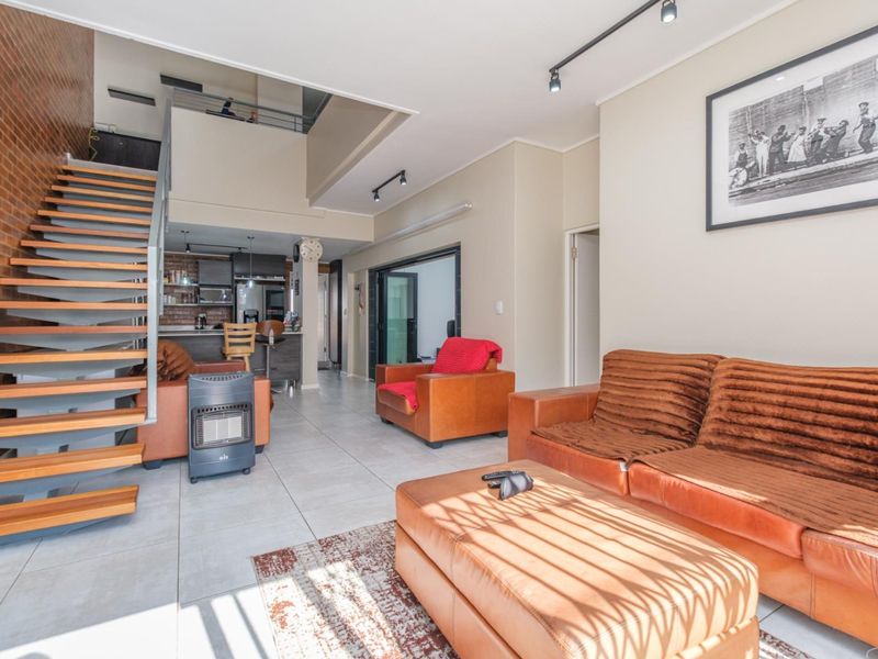 Spacious open-plan living area featuring an exposed brick wall, modern open-tread staircase, tiled flooring, and a mezzanine level overlooking the main living space.