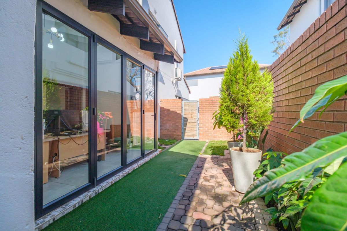 A well-maintained rear courtyard featuring a paved walkway, artificial lawn, brick boundary walls, and modern black-framed bi-fold doors leading into the home.