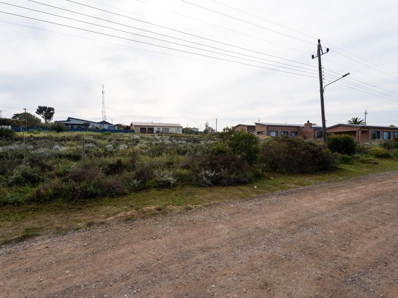A view of a vacant residential plot featuring natural vegetation, accessible via an unpaved road with nearby utility infrastructure.