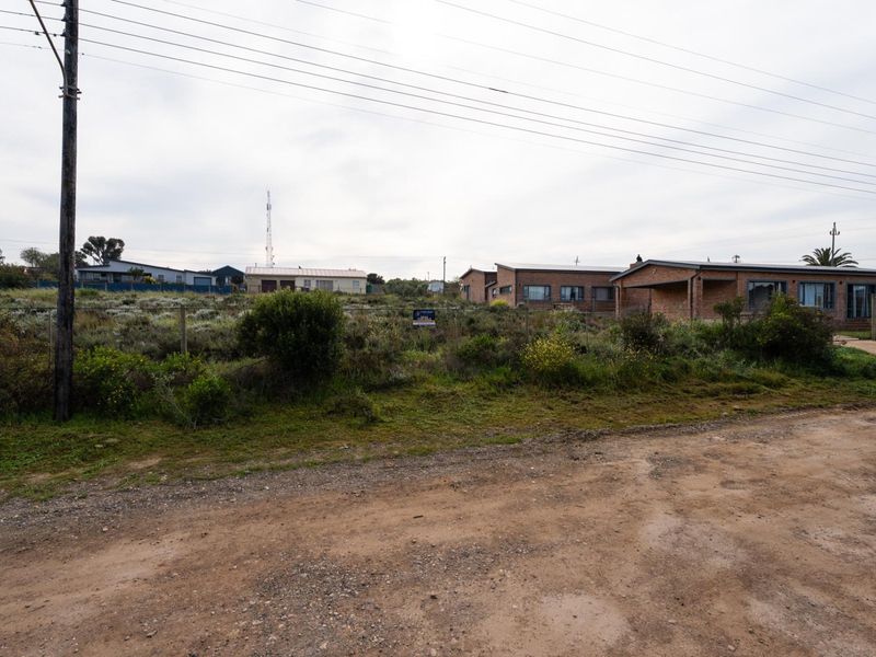 A street-level view of a vacant residential plot featuring an unpaved road, utility infrastructure, and surrounding neighborhood properties.