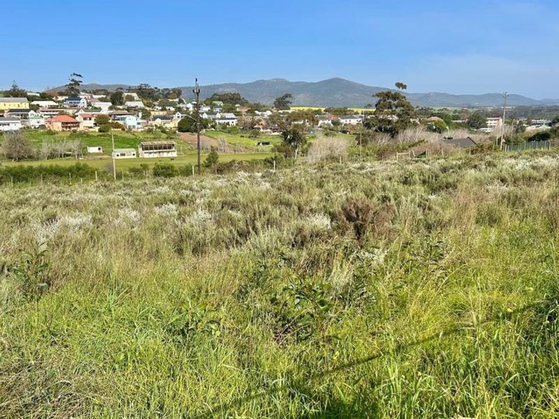A view of a vacant residential plot featuring natural vegetation, perimeter fencing, and a scenic outlook towards the surrounding town and mountain landscape.