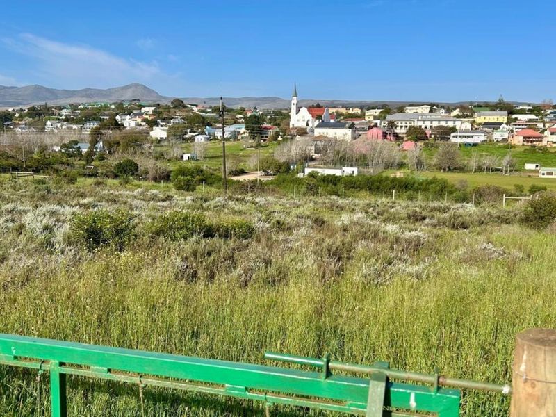 A scenic view overlooking a residential town with a church steeple, featuring a foreground of undeveloped land with perimeter fencing.