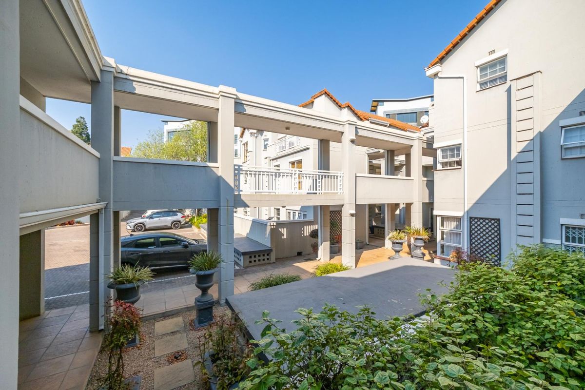 A view of the townhouse complex exterior featuring a covered walkway, paved courtyard area, and multi-level building architecture with terracotta roof tiles.
