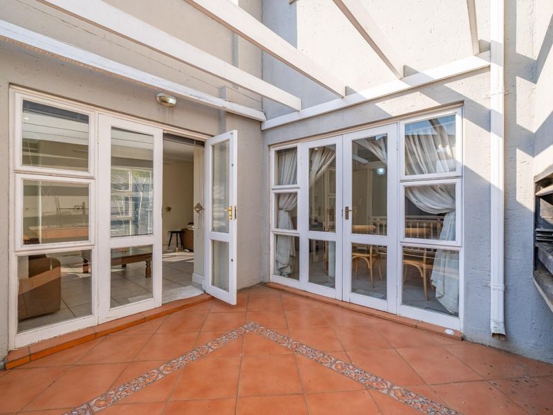 Covered patio area featuring terracotta tiled flooring, a built-in braai, and multiple sets of white-framed french doors providing access to the interior living spaces.