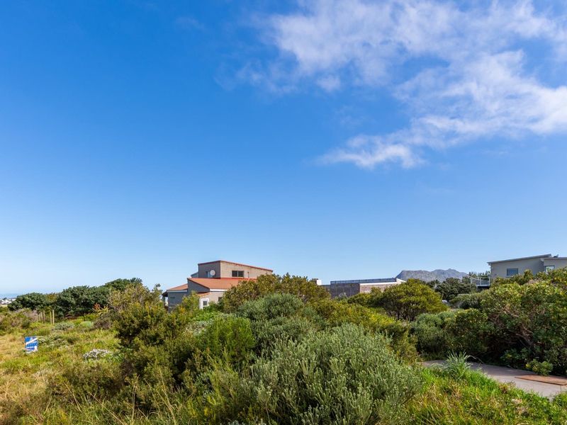 A view of a vacant residential plot featuring natural coastal vegetation and a dirt access path with surrounding neighborhood homes in the distance.
