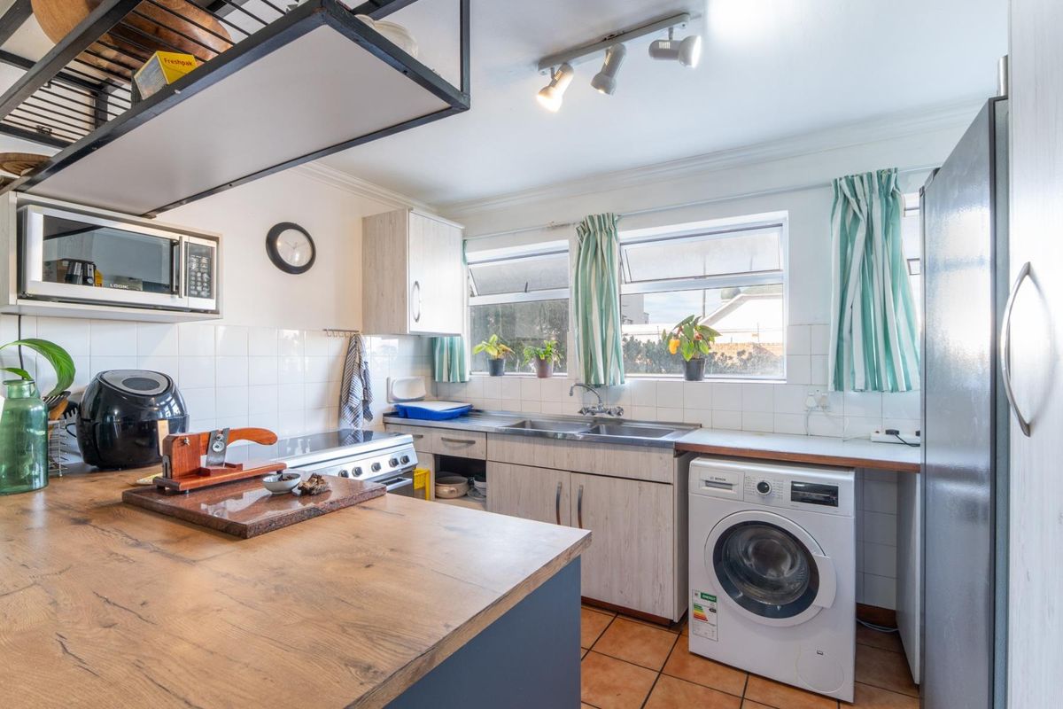 A functional kitchen featuring wood-look cabinetry, tiled backsplash, stainless steel sink, built-in oven, and integrated laundry appliance space with tiled flooring.