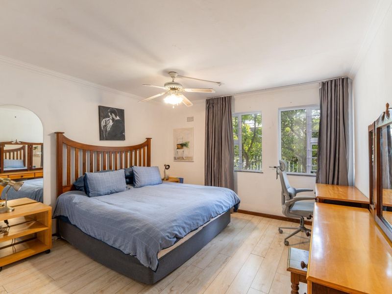 A well-lit bedroom featuring laminate flooring, a ceiling fan, and large windows that provide natural light and a view of the surrounding trees.