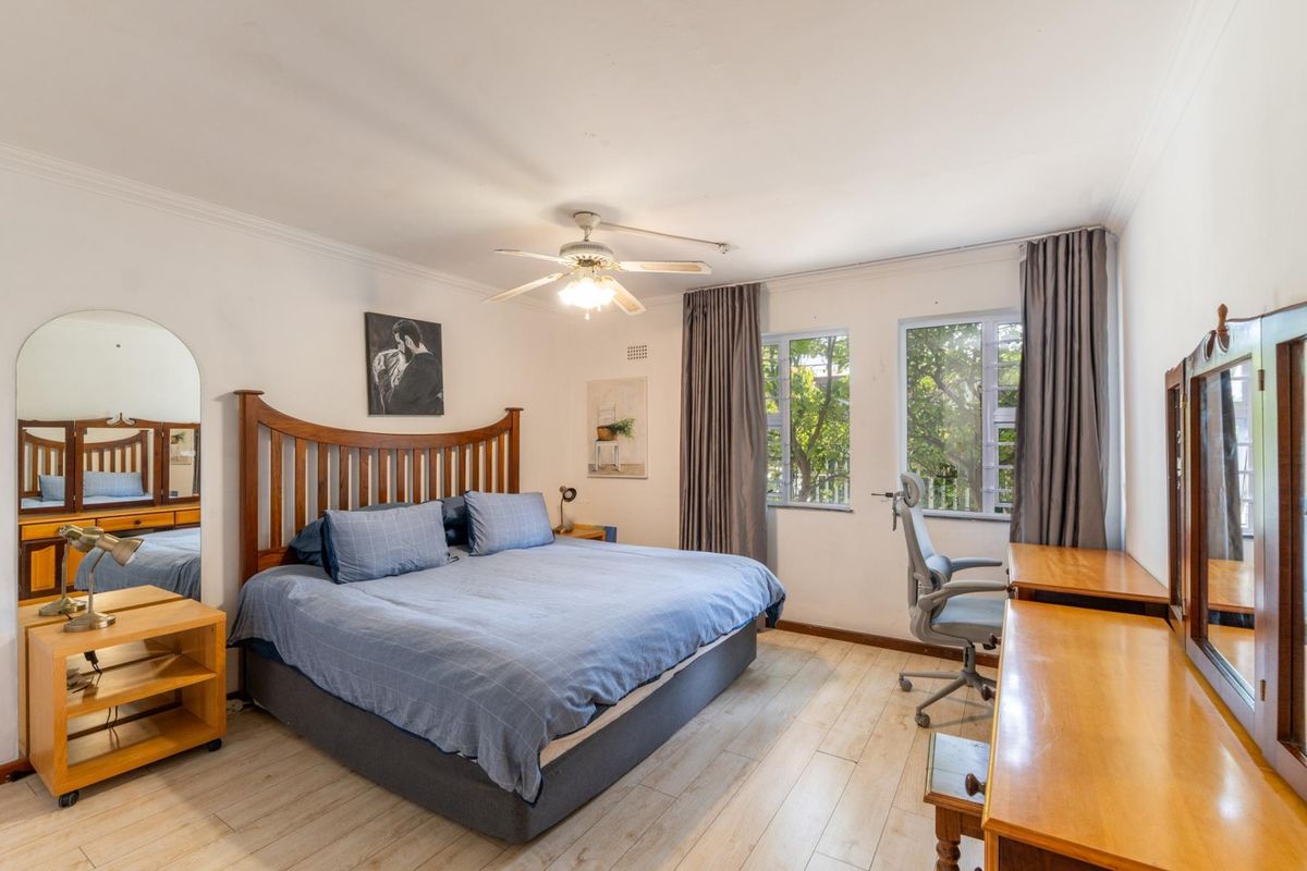 A well-lit bedroom featuring laminate flooring, a ceiling fan, and large windows that provide natural light and a view of the surrounding trees.