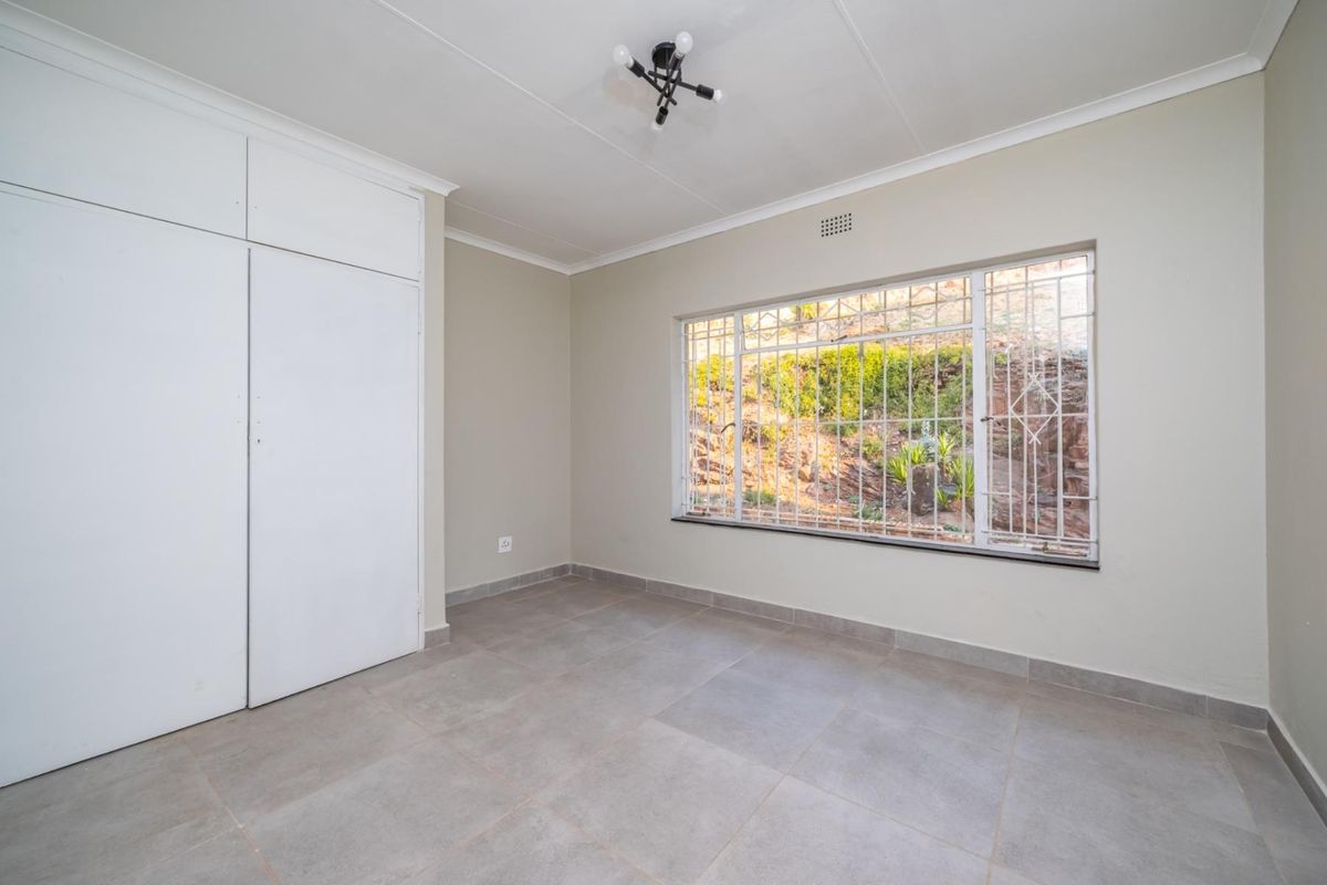 A bright, empty bedroom featuring neutral tiled flooring, built-in cupboards, and a large window fitted with security bars overlooking the garden.