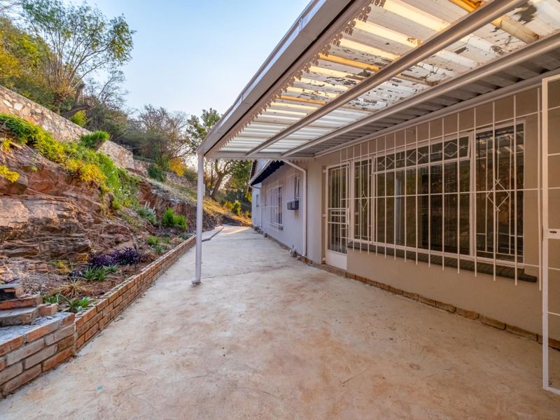 A covered outdoor patio area featuring concrete flooring, security bars on windows, and a corrugated roof, adjacent to a terraced garden.