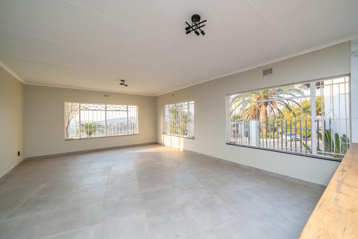 A spacious, empty living area featuring large windows with security bars, neutral tiled flooring, and modern ceiling lighting, offering a bright and versatile space.