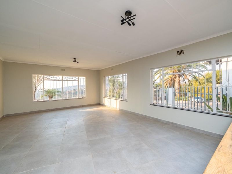A spacious, empty living area featuring large windows with security bars, neutral tiled flooring, and modern ceiling lighting, offering a bright and versatile space.