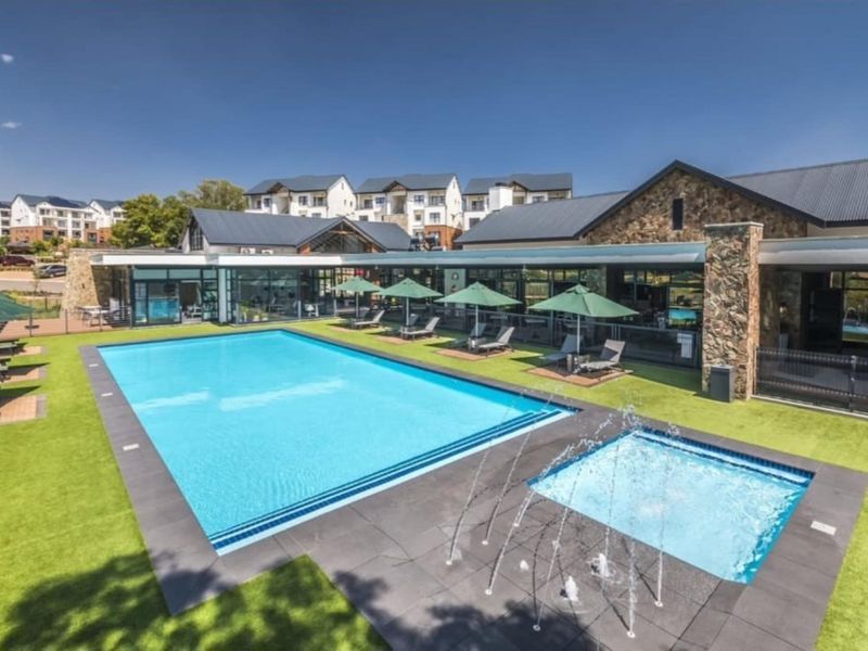 A well-maintained communal pool area featuring a large swimming pool, integrated water feature, stone-clad building exterior, and modern glass-fronted clubhouse facilities.