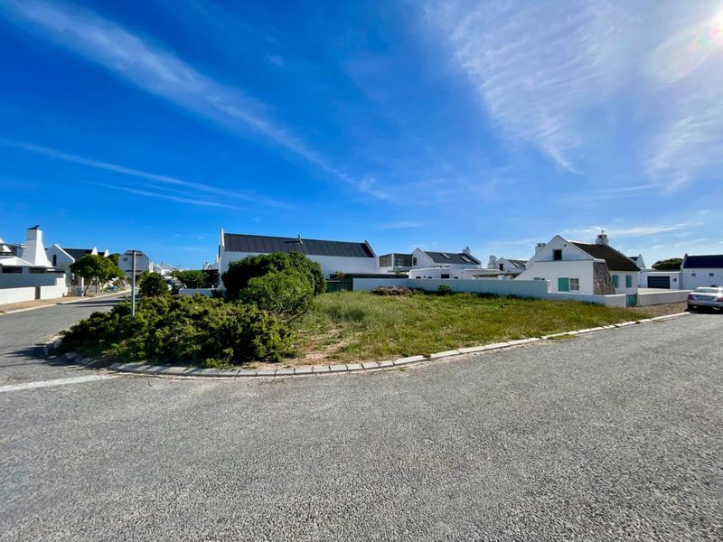 A street-level view of a residential corner plot featuring paved roads, concrete kerbing, and surrounding white-walled coastal style homes.