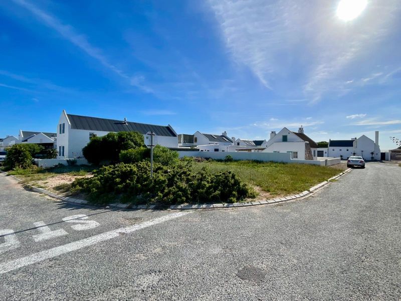 A street-level view of a residential corner lot featuring white-walled homes with dark metal roofing in a coastal estate setting.