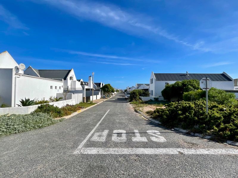 A clear street view of a residential estate featuring white-walled houses with dark roofs, well-maintained landscaping, and a paved road.