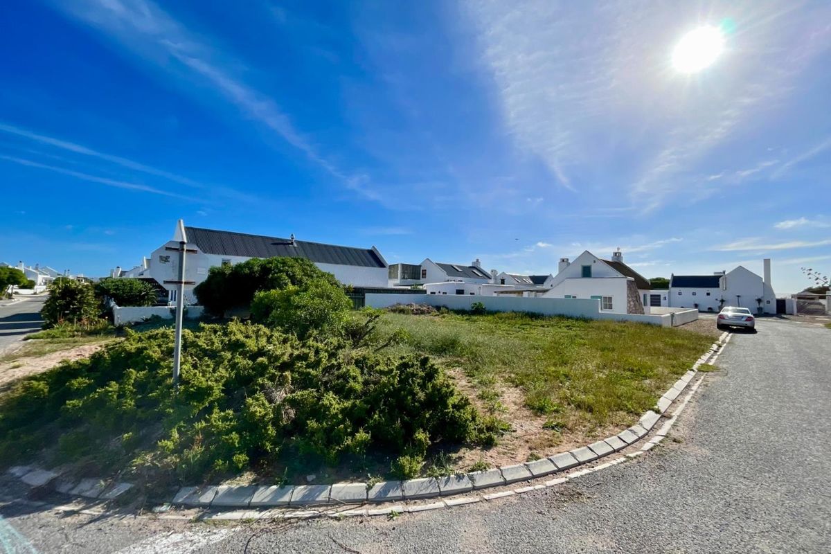 A street-level view of a corner plot featuring paved road access, concrete curbing, and surrounding residential properties in a coastal estate setting.