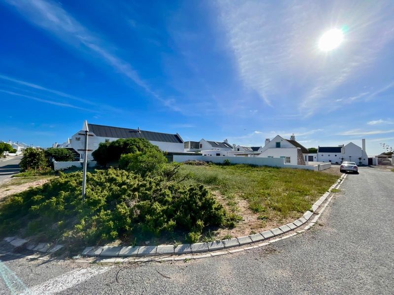 A street-level view of a corner plot featuring paved road access, concrete curbing, and surrounding residential properties in a coastal estate setting.