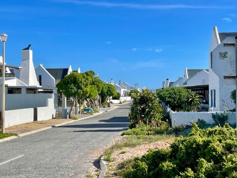 A clear street view of a residential neighbourhood featuring white-walled homes, paved roads, and established garden landscaping under a bright blue sky.