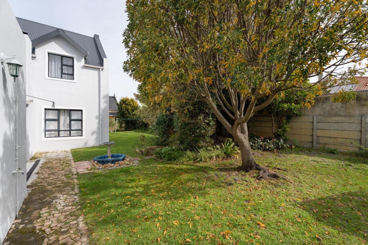 A view of the rear garden featuring a well-maintained lawn, mature trees, a cobblestone pathway, and a concrete boundary wall.