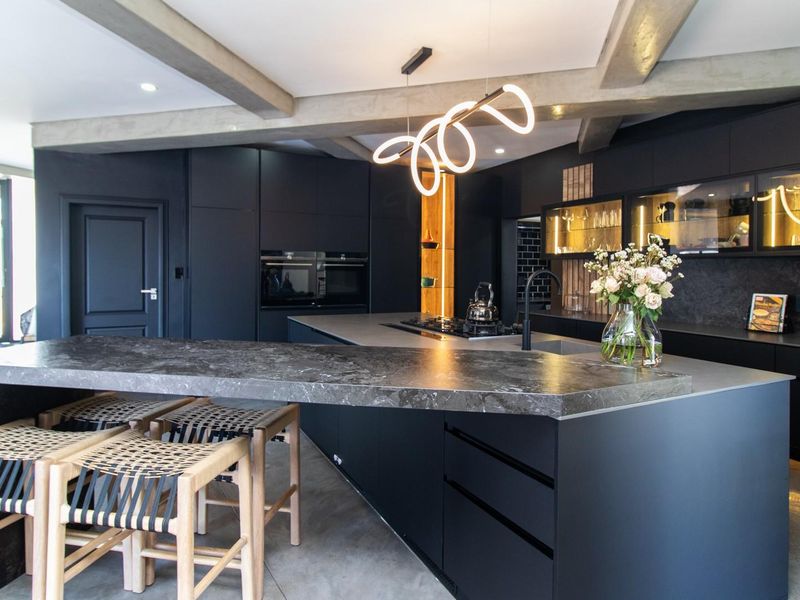 Modern, high-end kitchen featuring dark cabinetry, a large stone-topped island, integrated appliances, and striking exposed ceiling beams.