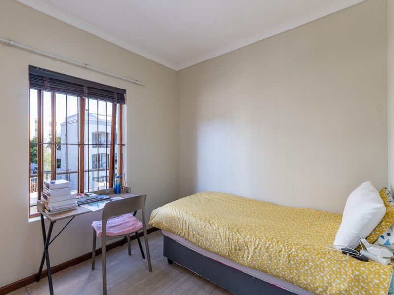 A compact bedroom featuring laminate flooring, neutral walls, and a window fitted with security bars and blinds.