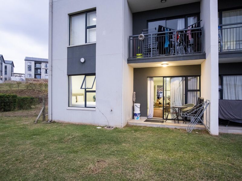 Rear view of an apartment building featuring a ground floor patio, upper balcony, and modern aluminum-framed windows with a small adjacent lawn area.
