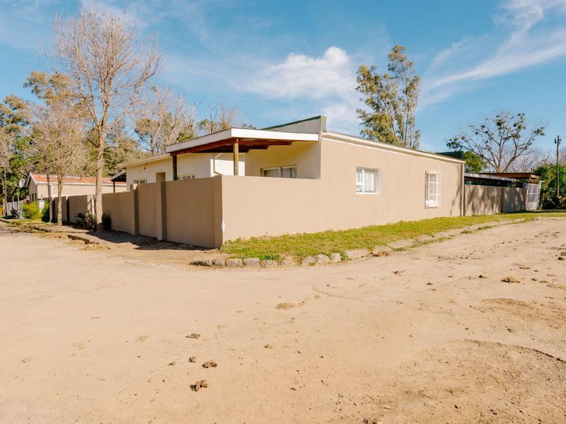 Exterior view of a single-story house featuring a perimeter wall, covered patio area, and sliding windows, situated on an unpaved road.