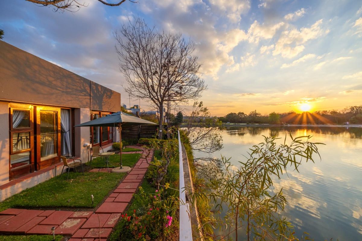 A scenic rear exterior view featuring a paved garden walkway, sliding glass doors, and a tranquil waterfront setting at sunset.