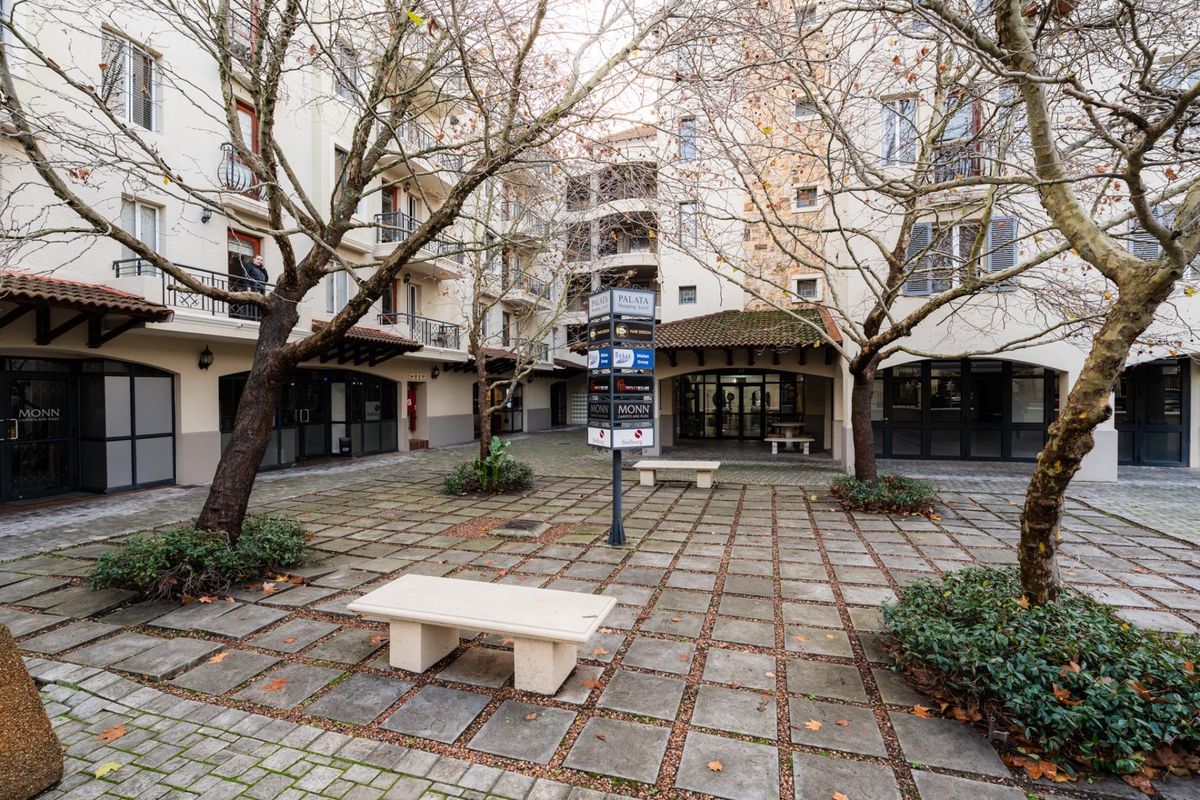 A paved courtyard area within an apartment complex, featuring stone benches, mature trees, and access to surrounding building units.