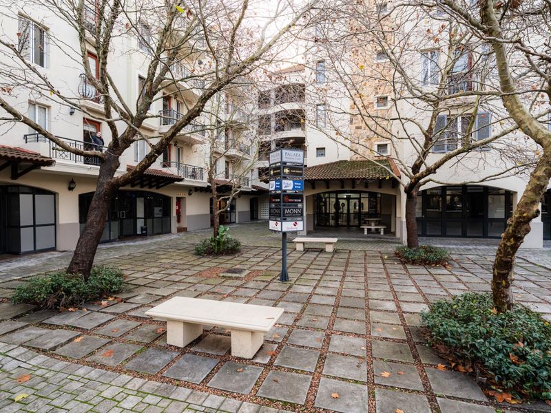 A paved courtyard area within an apartment complex, featuring stone benches, mature trees, and access to surrounding building units.