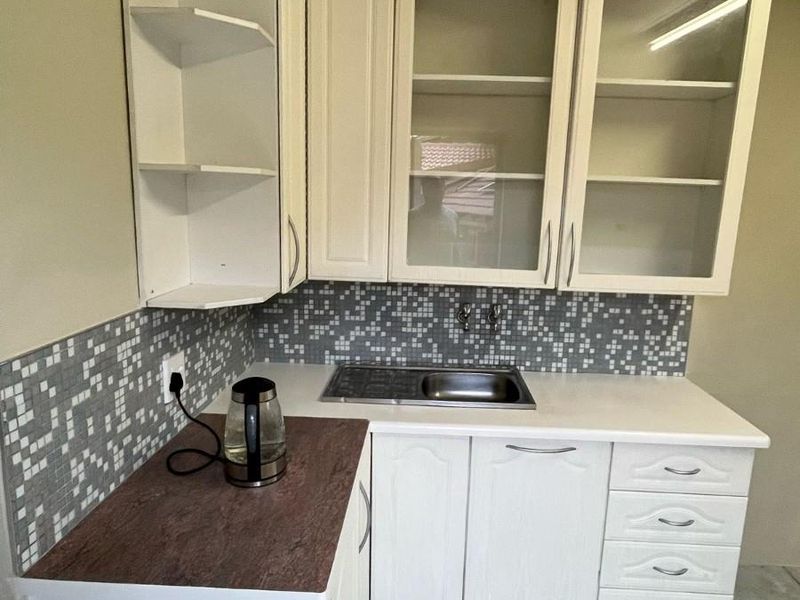 A compact kitchen area featuring white built-in cabinetry, a stainless steel sink, mosaic tile backsplash, and tiled flooring.